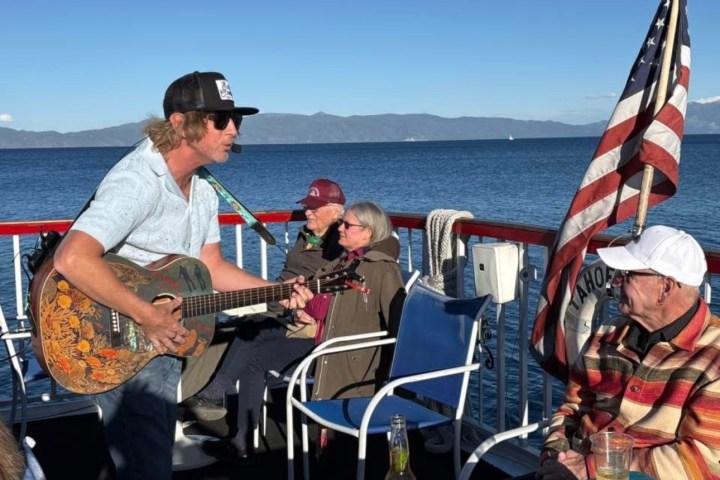 Man playing guitar on boat with seated people and U.S. flag, lake and mountains in background.