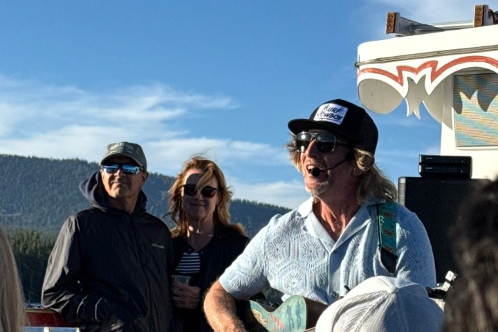 Person playing guitar and singing on a boat with scenic mountains and people watching.