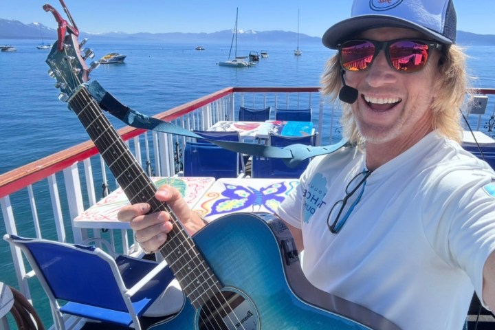 Person with guitar on boat, wearing hat and sunglasses, with water and boats in background.