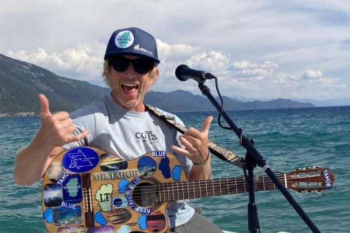 Man sitting on a boat, playing a sticker-covered guitar, with lake and mountains in the background.
